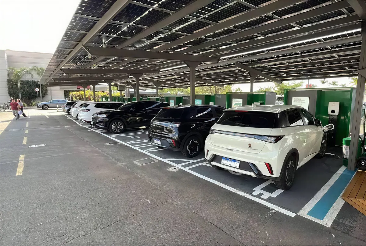 Carros estacionados nas vagas de recarga sob o carport solar, com a fileira de estações ao fundo