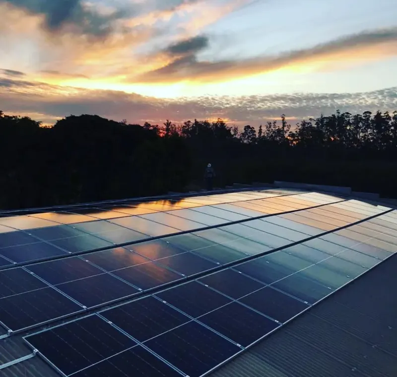 Usina solar no telhado metálico de um posto da rede Pica Pau ao entardecer, com a mata ao fundo