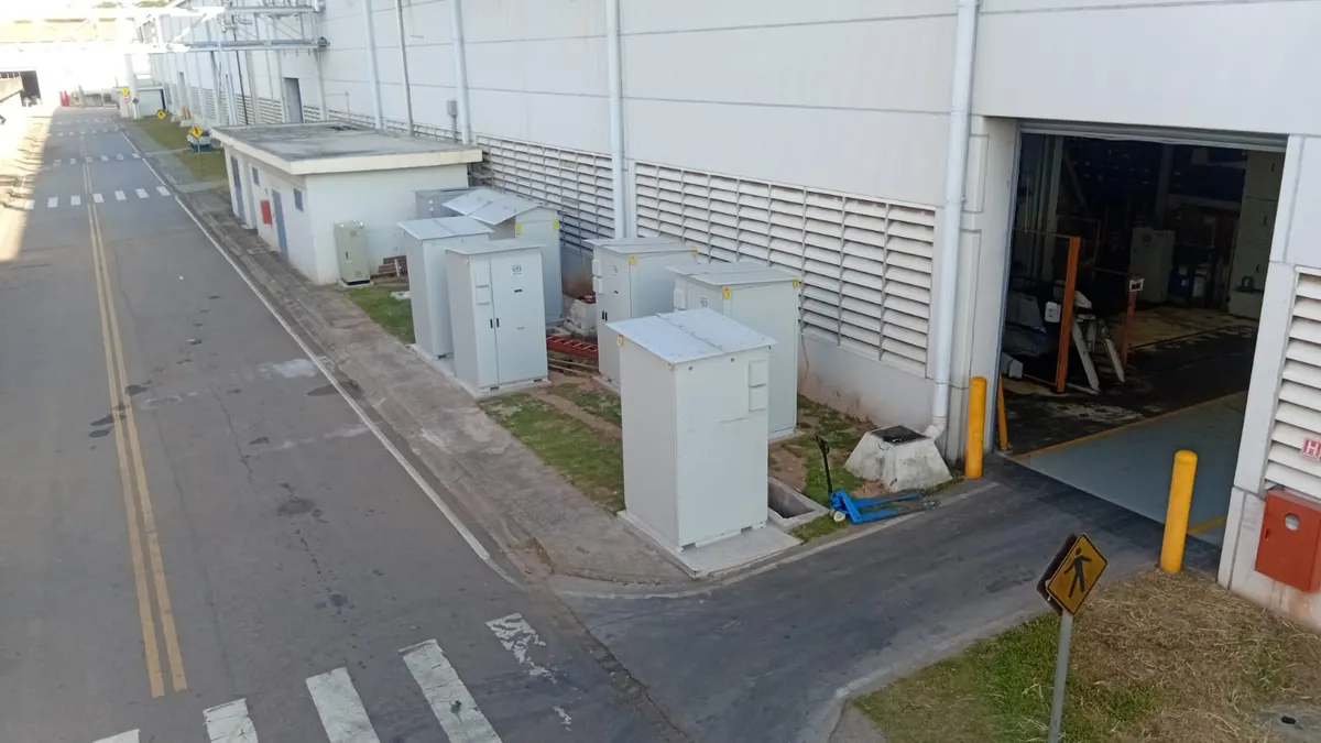 Sistema de baterias de uma indústria instalado ao lado do galpão, com os gabinetes alinhados junto à parede e a via interna da fábrica