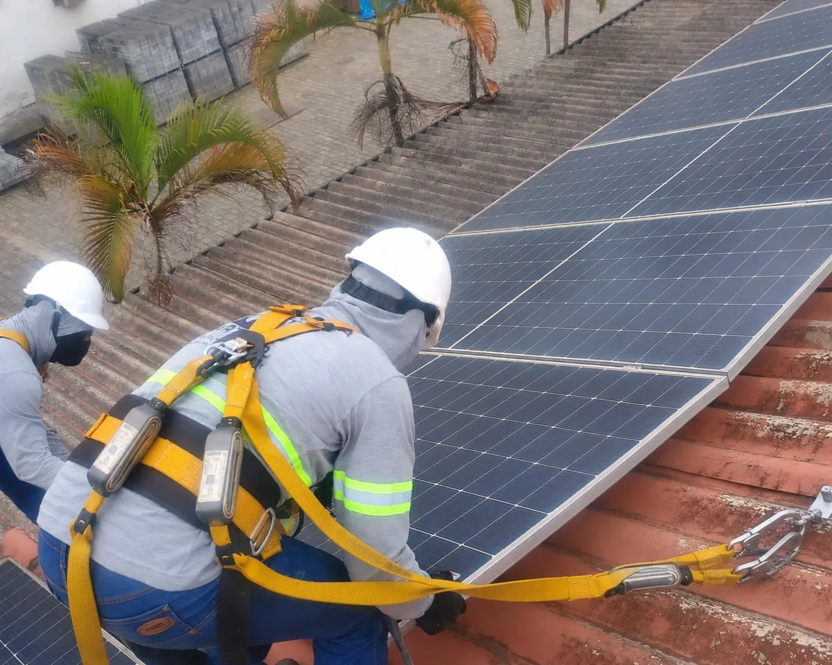 Dois técnicos de capacete, cinto paraquedista e talabarte trabalhando sobre um telhado de telha cerâmica com módulos solares