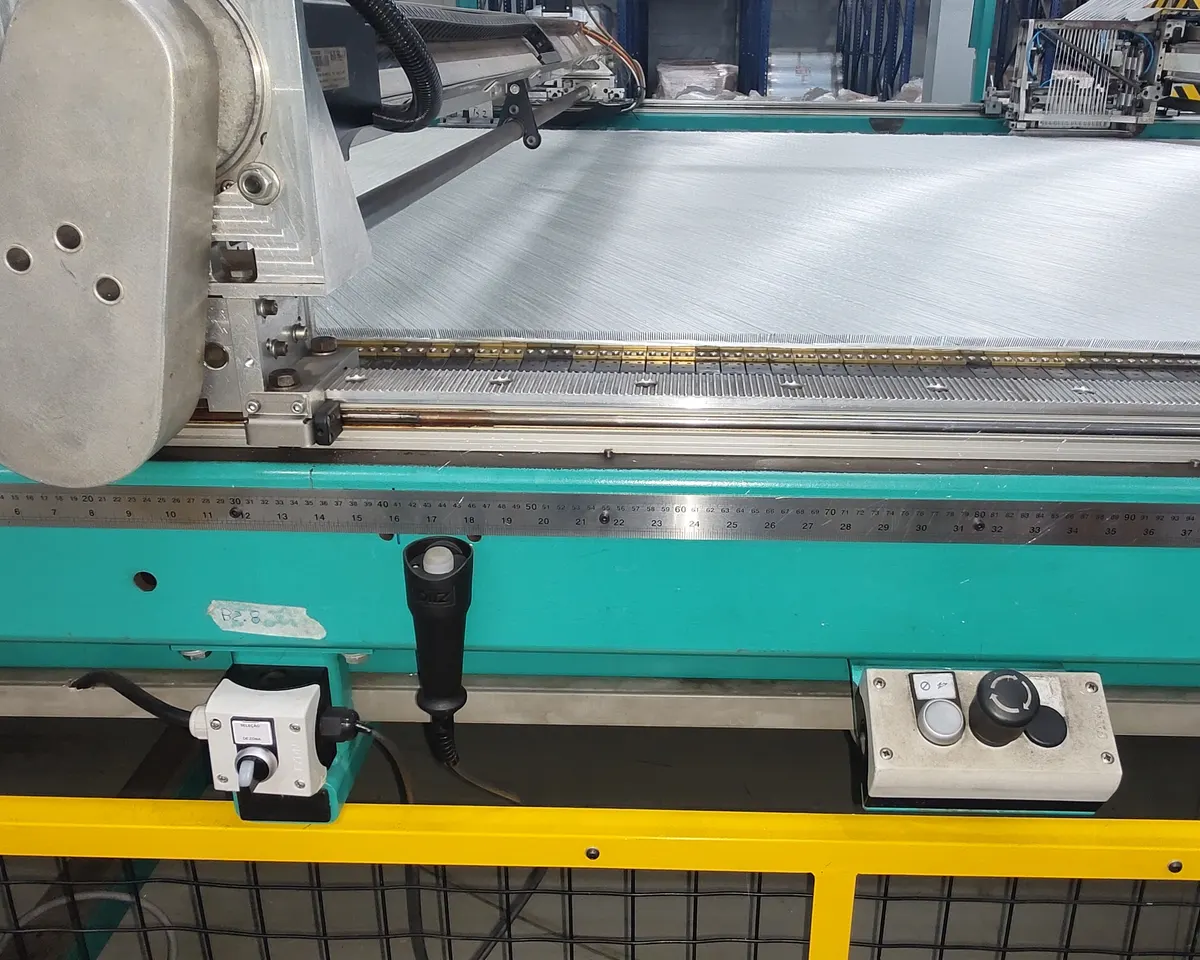 Mesa de corte industrial com botoeira de parada de emergência e comandos na lateral, e guarda-corpo amarelo com tela à frente