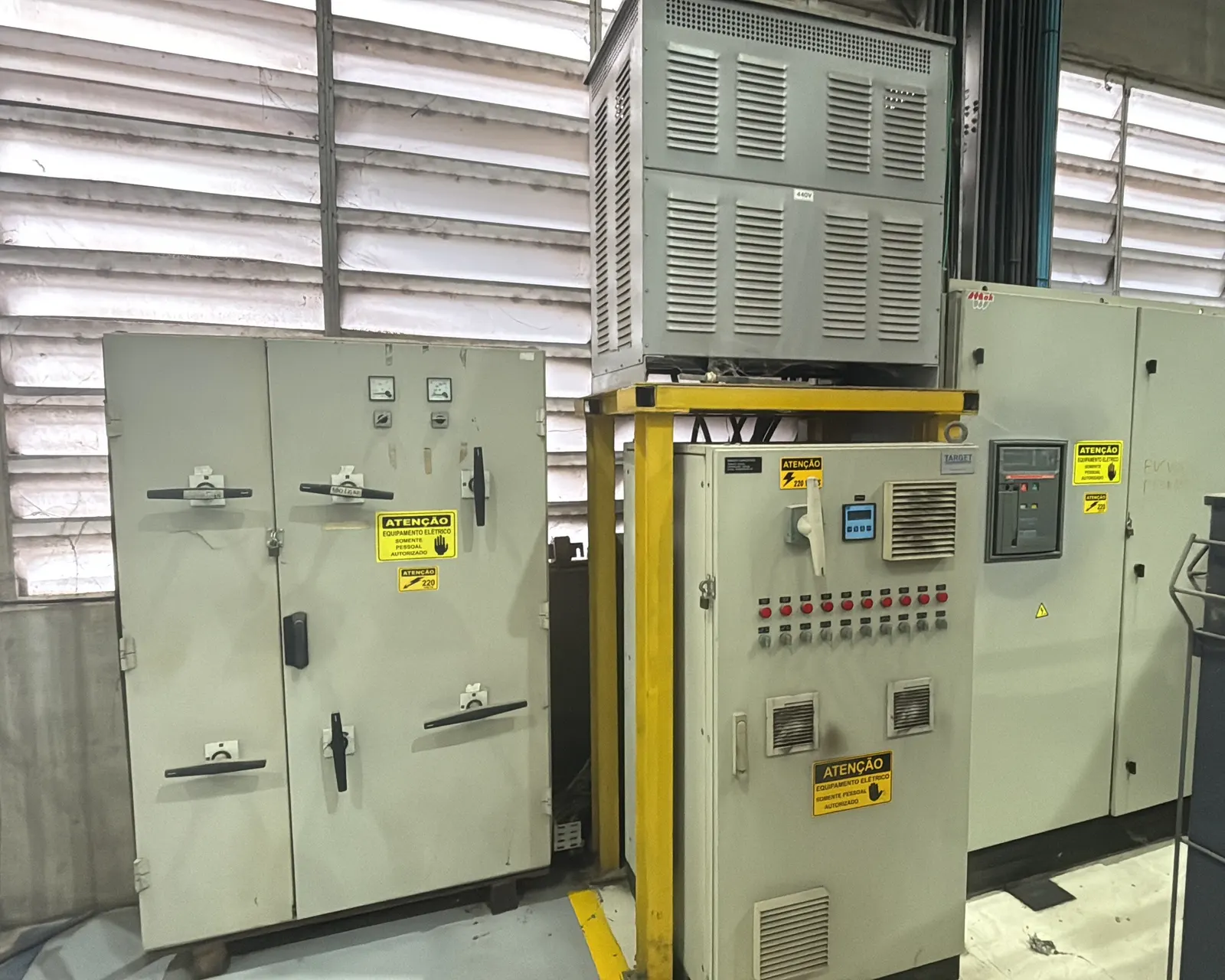 Quadro geral e centro de controle de motores de uma fábrica, com transformador a seco sobre estrutura amarela e etiquetas de atenção nos painéis